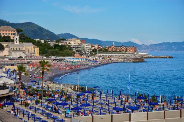 Une plage animée à Gênes, Italie, avec de nombreux parasols et chaises longues bleus, des bâtiments et des collines vertes à l'arrière-plan, et la mer bleue sur la droite.
