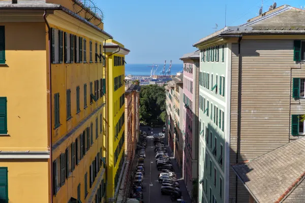 Une rue italienne colorée avec des bâtiments pastel, des voitures et une vue sur la mer et les grues du port.