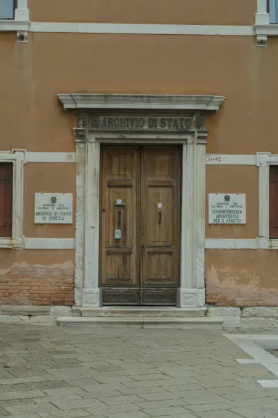 Une large porte ancienne en bois à l'entrée de l'Archivio di Stato à Venise, encadrée par des colonnes de pierre et une façade ocre.