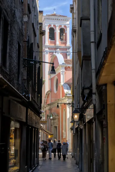 Photographie d'une ruelle étroite à Venise menant à un clocher rose avec des cloches et des arches, avec des gens qui passent.