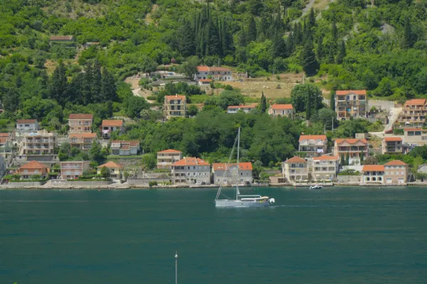 A sailboat on calm water in front of a coastal town with red roofs, nestled on a green hill.