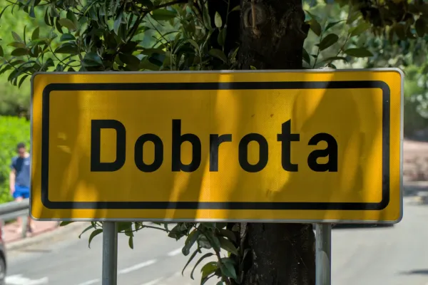 Señal amarilla con la inscripción 'Dobrota' en letras negras, rodeada de hojas verdes.
