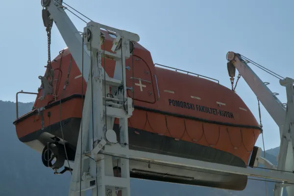 An orange lifeboat is suspended by a crane over the water against a blue sky.