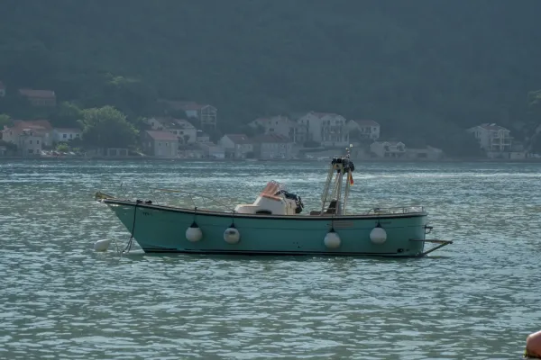 A turquoise boat with white fenders floats on calm waters in front of a coastal town in Kotor, Montenegro.