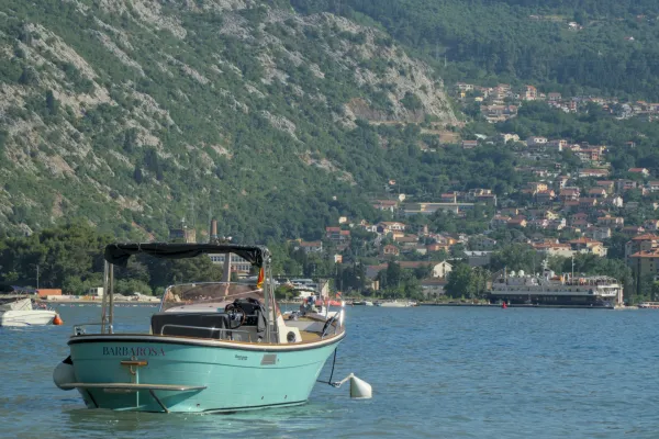 A turquoise boat rests by the coast of the Bay of Kotor, Montenegro, with green hills in the background.