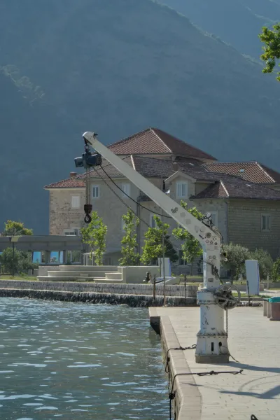Industrial crane on the shore of the Bay of Kotor, with a house and mountain in the background.