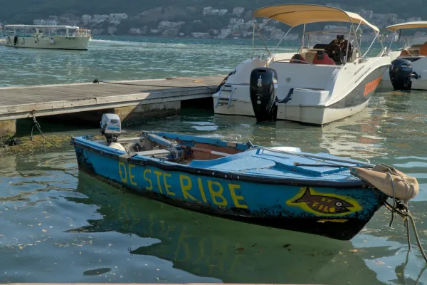 An old blue fishing boat with yellow lettering "DE STERIBE" and a fish symbol, floating in the Bay of Kotor near a pier and modern boats.