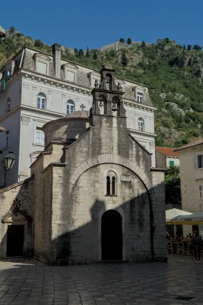Una antigua iglesia de piedra con campanario y fachada arqueada se alza ante una fortaleza montañosa en Kotor.