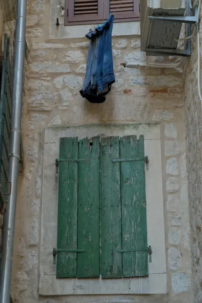 Window with green wooden shutters and jeans hanging to dry on a rustic stone wall in Kotor.