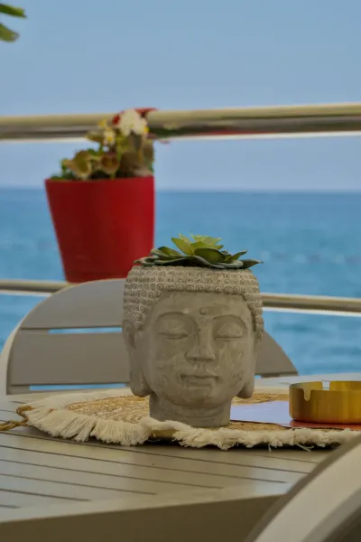 Una maceta con cabeza de Buda y una suculenta se encuentra sobre una mesa frente al mar azul.