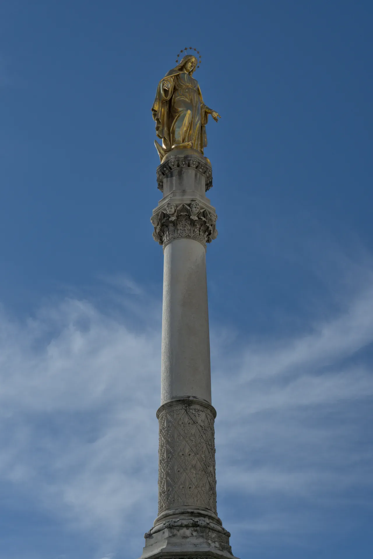 Zlatna statua Bogorodice na raskošno ukrašenom stubu na pozadini plavog neba sa blagim oblacima.