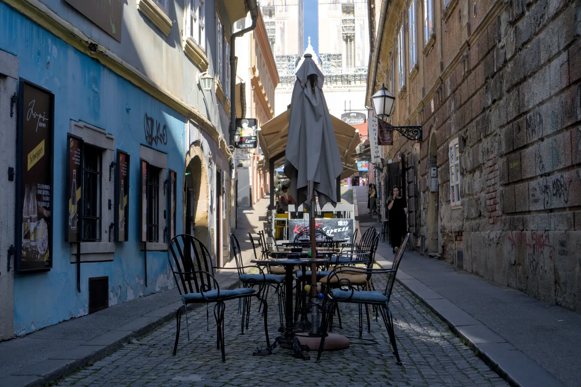 Uzak ulični prolaz u Zagrebu, sa zgradama plavih i kamenih fasada sa obe strane, i stolovima kafića u prvom planu.