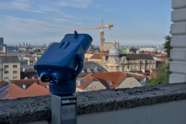 Un binocolo da osservazione blu si erge imponente sull'orizzonte pittoresco di Zagabria, con tetti rossi e una gru sullo sfondo.