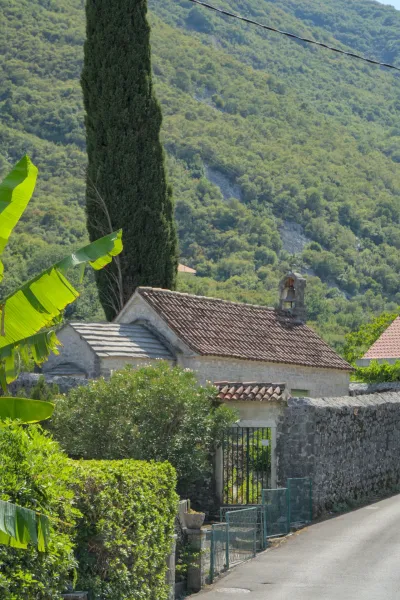 Small stone church with terracotta tiled roofs, surrounded by lush greenery and a tall cypress tree, near Kotor, Montenegro.