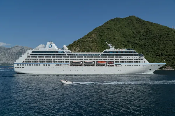 A large white cruise ship leaving Kotor Bay, with a small boat alongside.