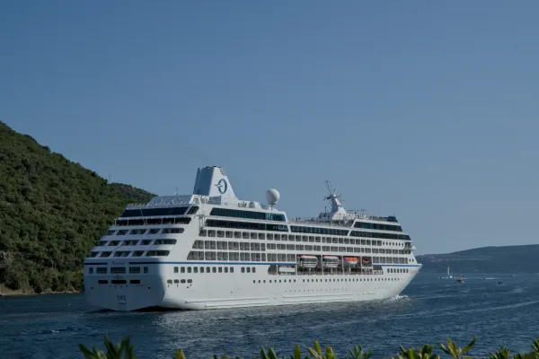 Un gran crucero blanco parte de la bahía de Kotor bajo un cielo azul despejado, con las colinas verdes de Montenegro al fondo.