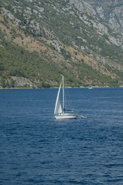 A small sailing yacht floats on the water near Kotor, Montenegro, with steep, green-covered mountains in the background.