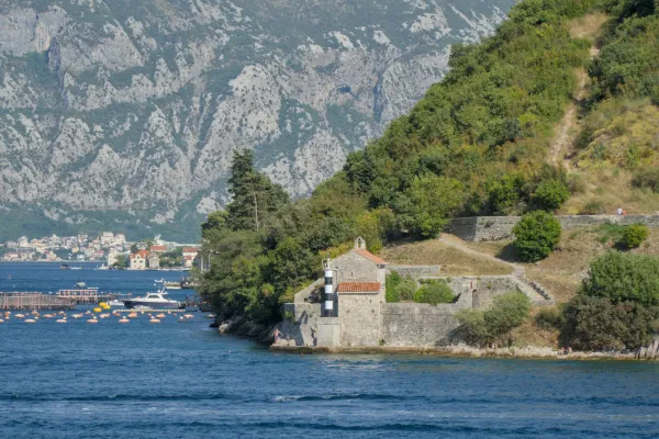 A small stone chapel with a black and white striped lighthouse on the shore, backed by mountains and blue water in Kotor.