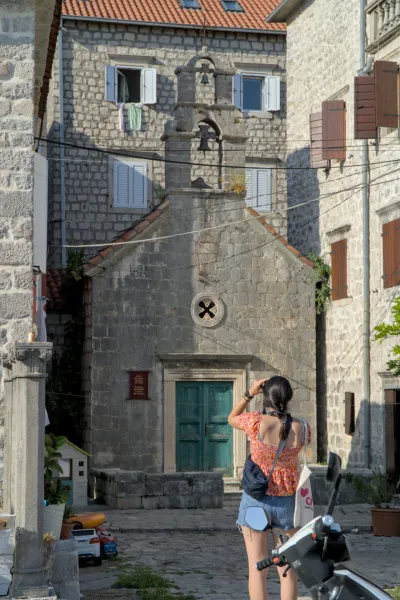 A woman photographs the "Perast Alley" in Kotor, Montenegro, featuring a stone church facade and Venetian architecture.