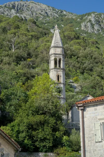 A stone bell tower rises amidst lush greenery, with rocky slopes in the background, on a clear evening in Perast.