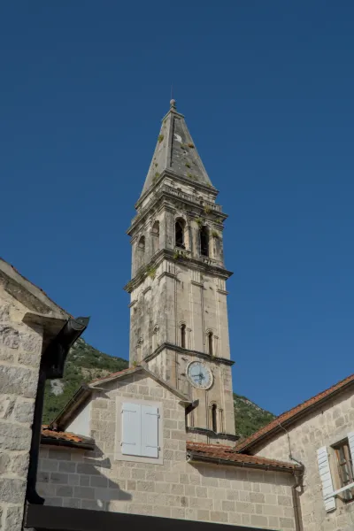 Famous bell tower of Perast, Montenegro, under a bright blue sky, with stone buildings in the foreground.