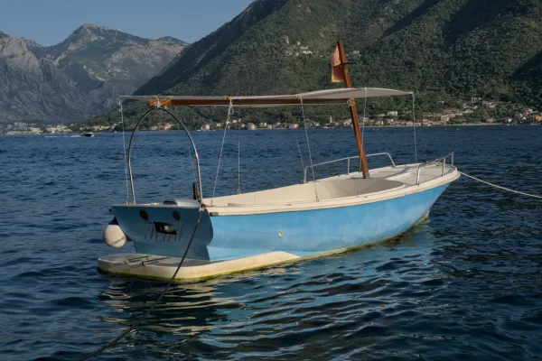 A blue boat is moored in the water off the coast of Perast, Montenegro, with mountains and houses in the background.