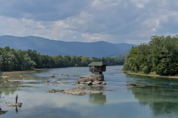 A small house on a rock in the Drina River, with a lone fisherman casting his fishing rod nearby.