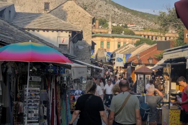 Оживленная улица Старого города Мостара в сумерках, с магазинами и горами на заднем плане.