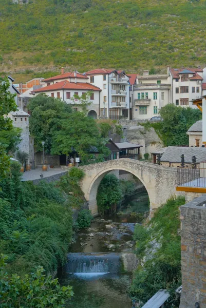 "Puente Torcido" en Mostar, Bosnia y Herzegovina, con una pequeña cascada y abundante vegetación, en formato vertical.