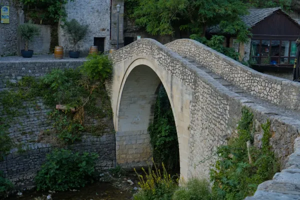 Un puente de piedra de un solo arco conocido como el 'Puente Torcido' o Kriva Ćuprija en Mostar, Bosnia y Herzegovina, rodeado de vegetación.