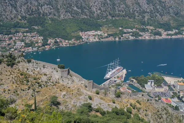 A huge sailing ship is docked in Kotor port, Montenegro, with ancient fortress walls rising above it.