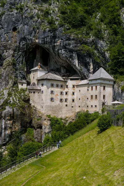 Château de Predjama construit dans une paroi rocheuse massive, avec une végétation verte et des visiteurs sur un chemin en contrebas.