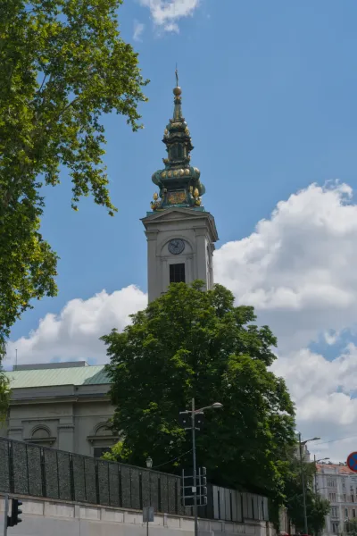 Eleganter Kirchturm in Belgrad vor blauem Himmel mit Wolken, umrahmt von Bäumen.