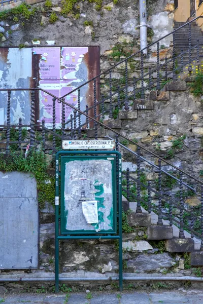 Eine alte Steintreppe mit einem verzierten Metallgeländer führt neben einer Steinmauer nach oben. Im Vordergrund zeigt eine grüne Anschlagtafel verwitterte Poster, und überwucherte Pflanzen klettern die Wand und die Treppe hoch.