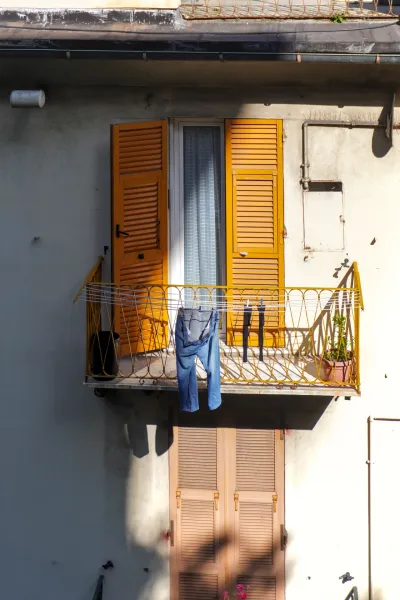 Un balcon à Gênes, Italie, avec des volets orange vif sur la fenêtre, un jean bleu et des chaussettes noires sur une corde à linge, et une petite plante verte en pot.