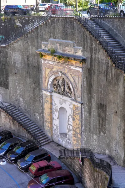 Un monument historique patiné avec un relief sculptural sous une arche, adossé à un mur de pierre rugueux, comportant de nombreux escaliers extérieurs et un parking avec des voitures en contrebas à Gênes, Italie.