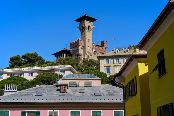 Pogled na Castello Mackenzie, zamak u Đenovi, Italija, sa svojom upečatljivom kulom, na pozadini zelenog drveća i stambenih zgrada pod jarko plavim nebom.