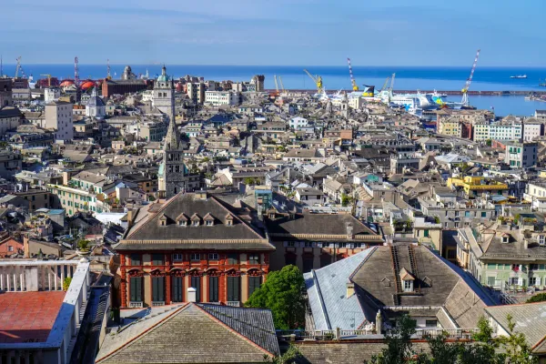 Eine Weitwinkelaufnahme von Genua, Italien, die den aktiven Hafen mit Frachtschiffen, Fähren und Kränen zeigt, mit Blick auf die dichte städtische Landschaft, die sich bis zum blauen Meer erstreckt.