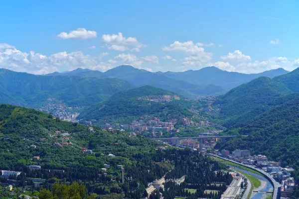 Blick auf grüne, baumbestandene Hügel in der Nähe von Genua, Italien, mit alten Befestigungsanlagen und modernen Kommunikationstürmen auf den Gipfeln unter strahlend blauem Himmel.