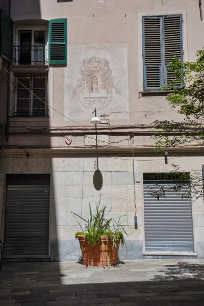 Une rue étroite et ensoleillée à Gênes, Italie, avec des bâtiments présentant des volets verts et bleus, une fresque délavée sur le mur représentant une scène religieuse, un seul lampadaire, un grand pot en terre cuite avec des plantes et deux portes roulantes de magasins projetant des ombres sur le sol pavé.