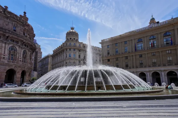 Velika fontana na više nivoa koja prska vodu visoko u vazduh u centru Piazza De Ferrari u Đenovi, Italija, sa klasičnim zgradama i plavim nebom u pozadini.