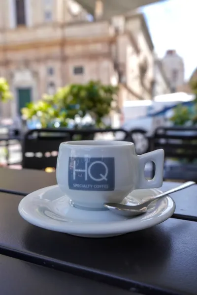 Белая эспрессо-чашка с логотипом 'HQ Specialty Coffee' и ложкой на блюдце, на черном столе в уличном кафе в Генуе, Италия. Фон размыт, видны здания и зелень.