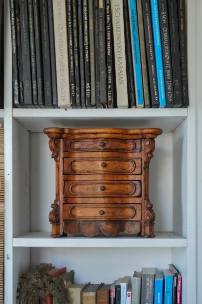 Une vue rapprochée d'une petite commode ancienne en bois avec cinq tiroirs, chacun avec un petit bouton rond, posée sur une étagère blanche. Au-dessus de la commode, une rangée de boîtiers de CD avec des titres de musique classique est visible. Sous la commode, une autre étagère contient une variété de livres et une pièce métallique décorative.