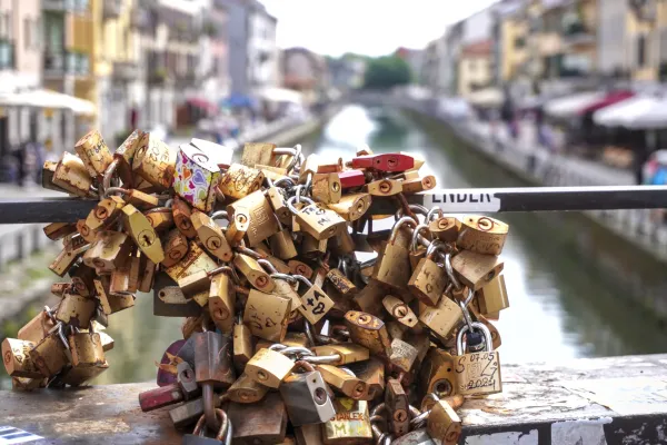 A pile of weathered and colorful padlocks attached to chains on a bridge railing, with a blurred canal and buildings of Milan in the background.