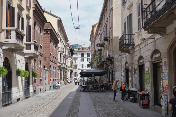 Ein Mann auf einem Fahrrad überquert eine Fußgängerzone in Mailand, Italien. Entlang der Gasse befinden sich Gebäude mit Fensterläden, Balkonen, Graffiti und Geschäften.