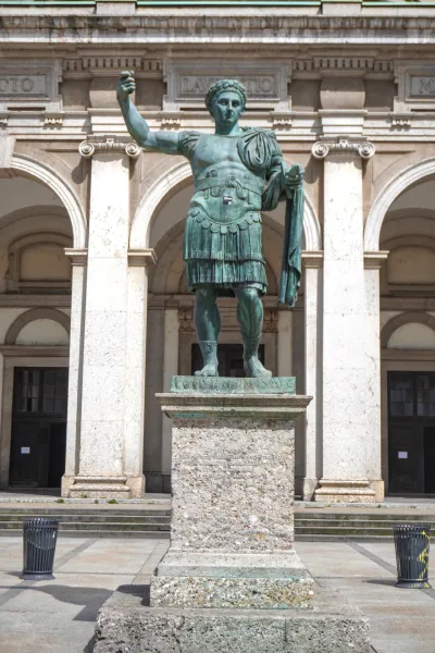 A tall bronze statue of Emperor Constantine the Great in military uniform, standing on a stone pedestal in front of a building with grand arches and classical architecture in Milan, Italy.