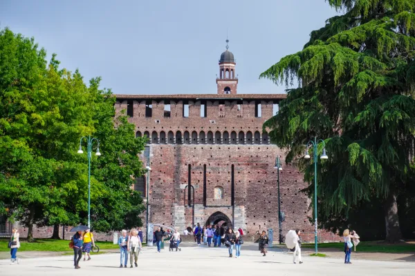 Persone passeggiano davanti al Castello Sforzesco a Milano, Italia, con grandi alberi che incorniciano la vista.