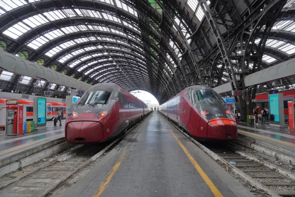 Two modern red high-speed trains wait on the platforms at Milan Central Station under its grand arched glass roof.