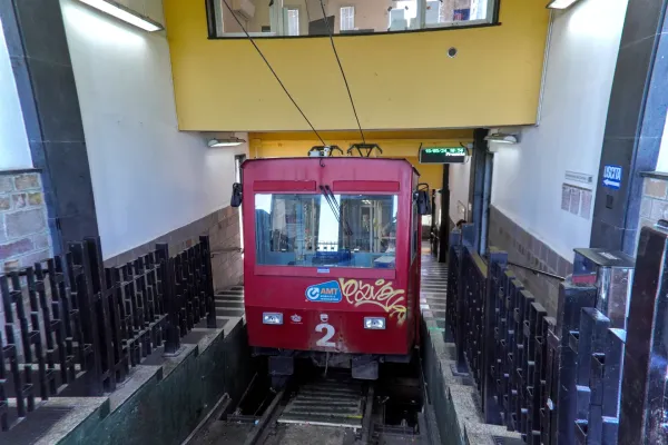 Un train de funiculaire rouge est garé dans une gare avec des escaliers menant à la sortie et des portillons de billetterie au premier plan.