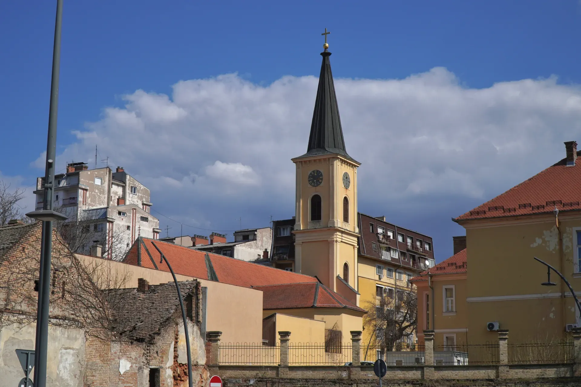 Una torre de iglesia amarilla con un techo puntiagudo negro y un reloj es visible en Pančevo, Serbia. Se ven edificios modernos en el fondo, y el cielo es azul con nubes blancas.